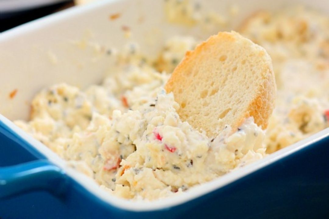 A slice of bread is dipped into a creamy cheese spread with herbs and diced vegetables in a blue rectangular dish.