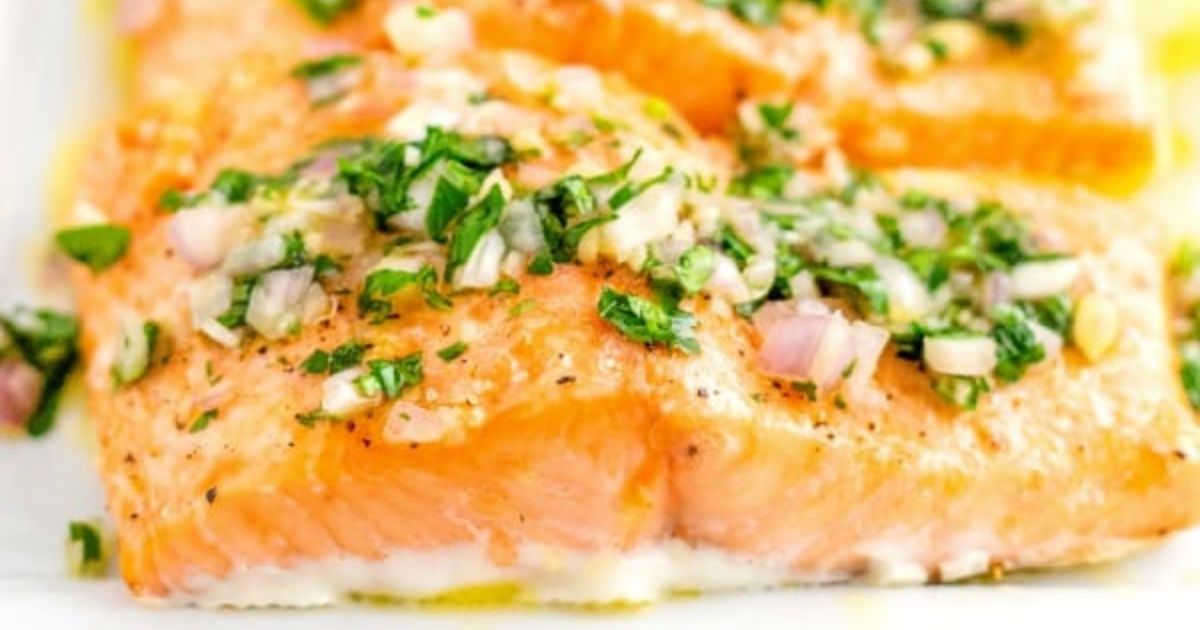 Close-up of cooked salmon fillet topped with chopped herbs, onions, and a light drizzle of oil on a white plate.