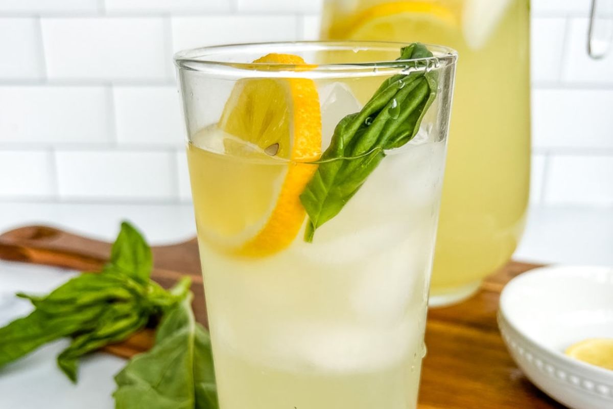 A glass of iced lemonade garnished with lemon slices and fresh basil sits on a wooden surface, with a pitcher and more basil leaves in the background.