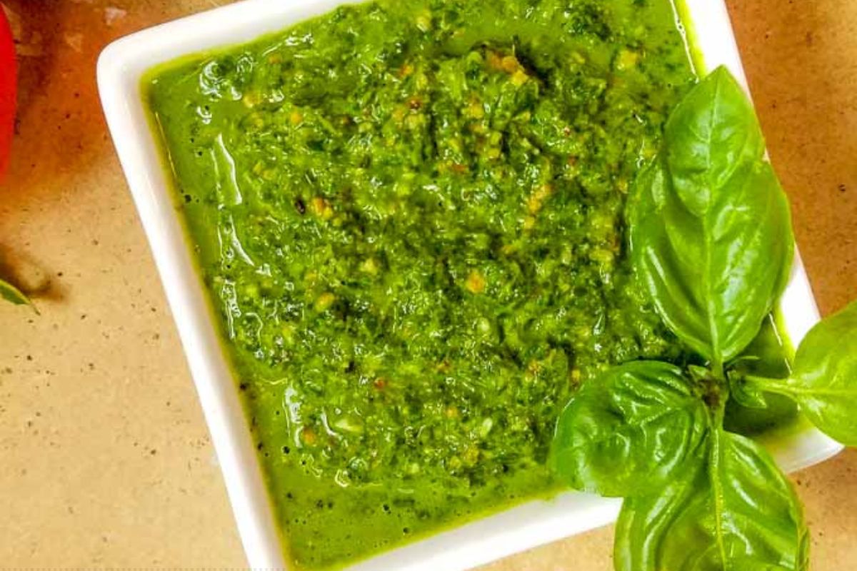 Square white bowl filled with green pesto sauce, garnished with fresh basil leaves, on a tan countertop