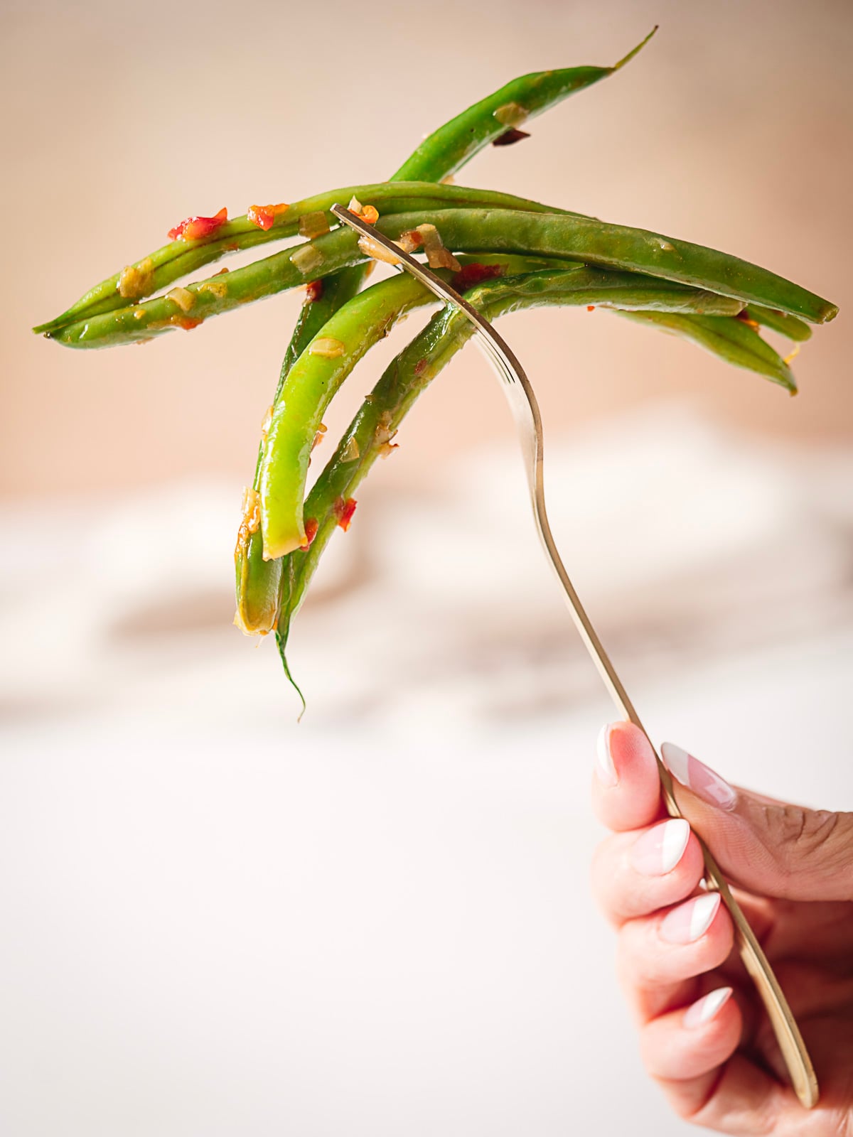 A hand holding a fork with cooked green beans topped with small bits of seasoning and chili.