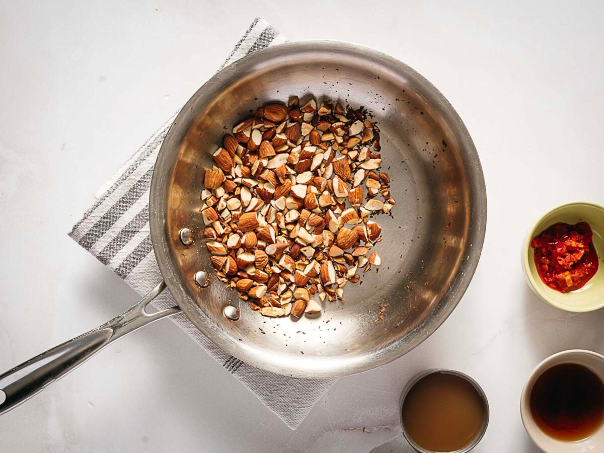 A stainless steel pan with chopped almonds sits on a striped towel, surrounded by small bowls containing red peppers, a brown liquid, and a dark sauce.