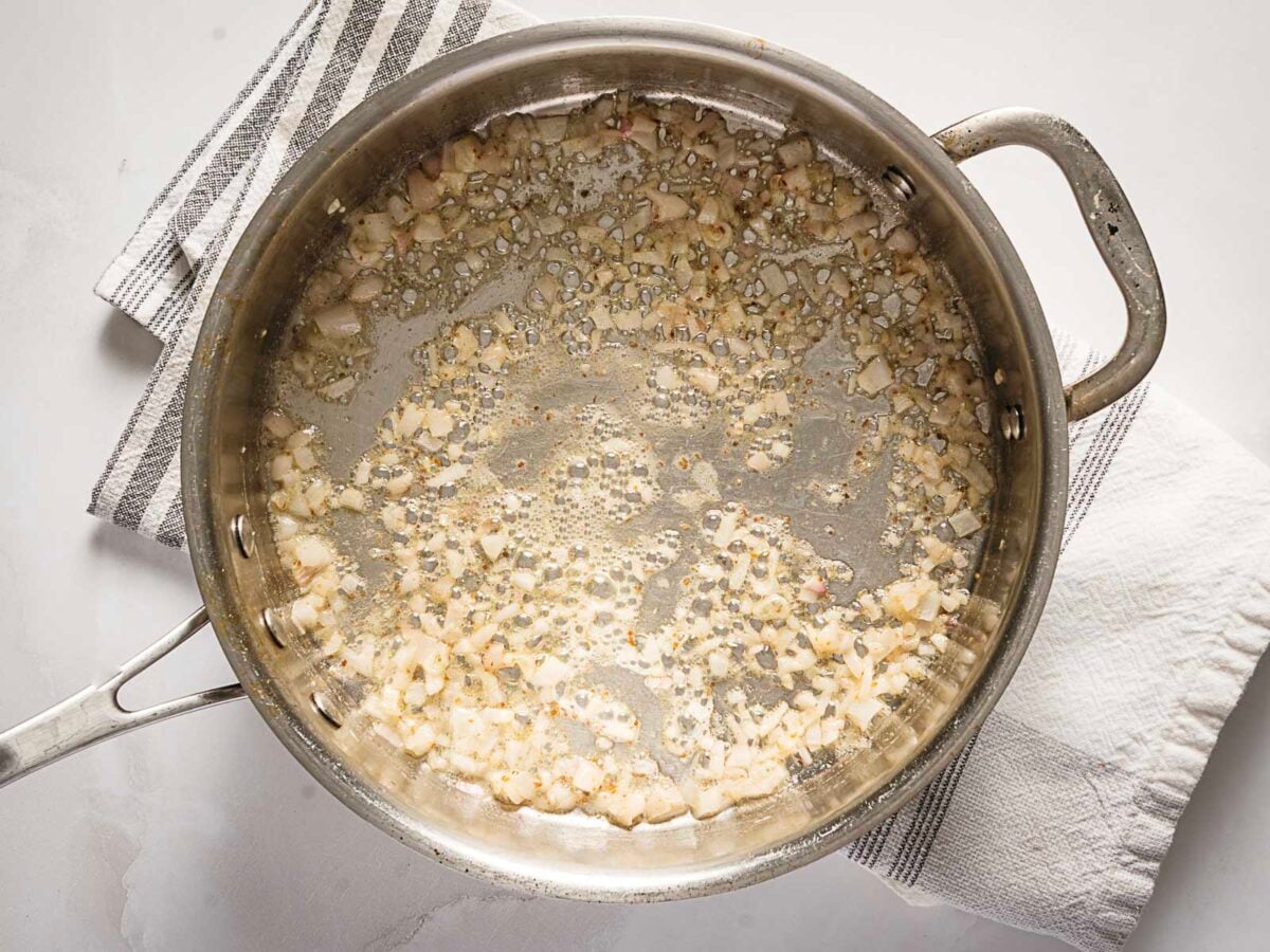 Diced onions are being sautéed in oil or butter in a stainless steel skillet. The pan is on a white surface with striped and white kitchen towels beside it.