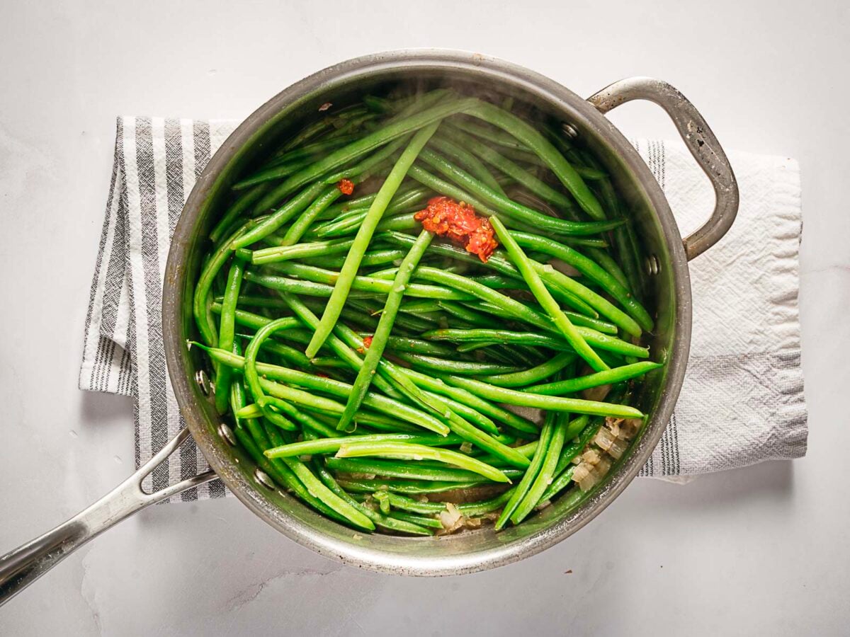 A stainless steel pan filled with cooked green beans and a small amount of red seasoning, sitting on a striped kitchen towel.