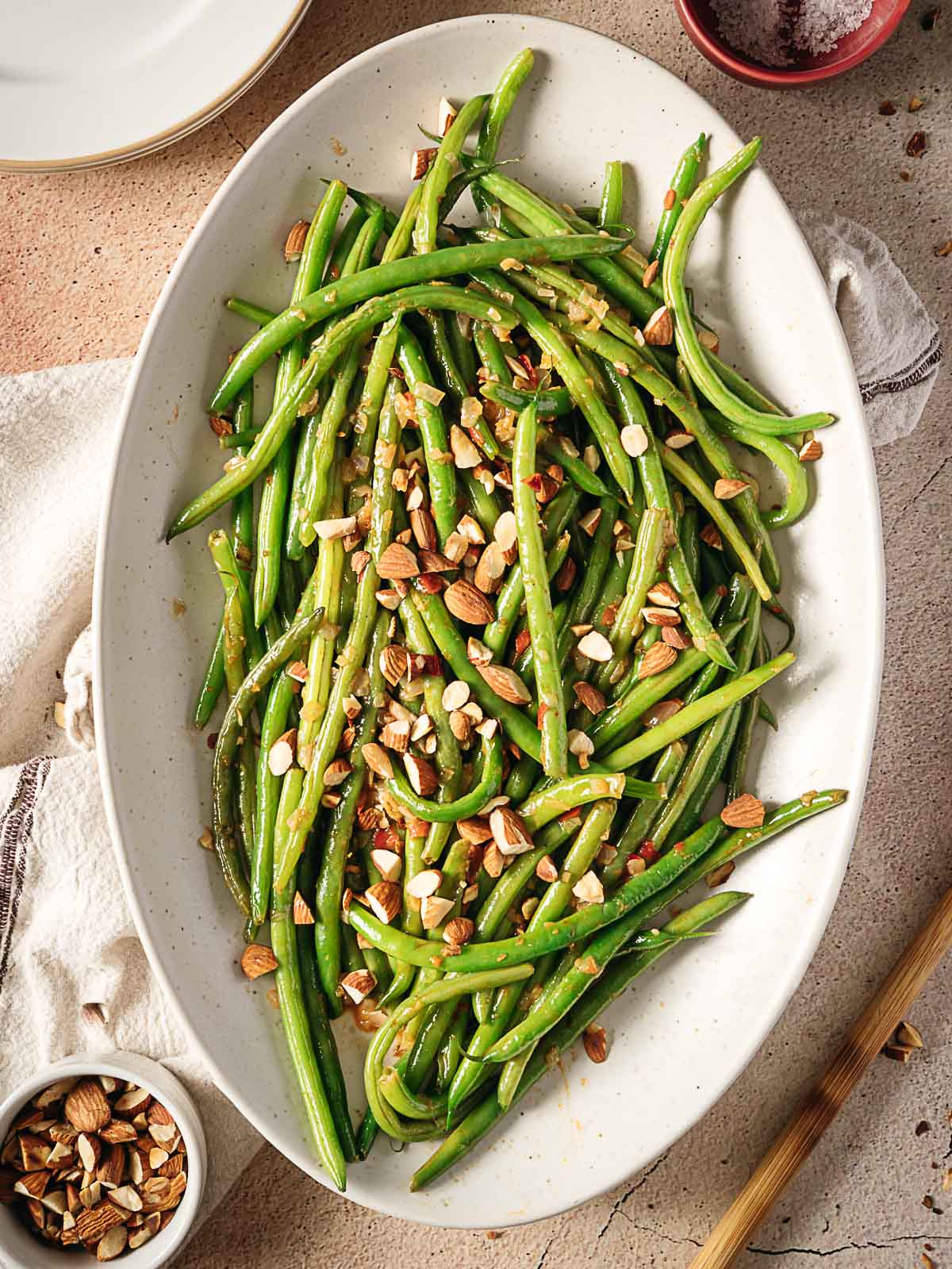 Oval platter with cooked green beans topped with chopped almonds and crumbled cheese, placed on a neutral surface with small bowls of almonds and salt nearby.
