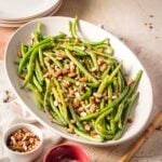 Oval platter of cooked green beans topped with chopped almonds, set on a table with a small bowl of almonds and plates nearby.