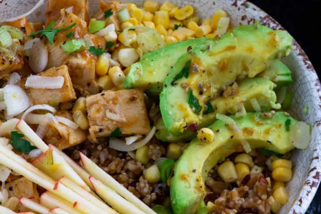 A close-up of a bowl containing sliced avocado, diced tofu, corn, apple slices, and grains, garnished with chopped onions and herbs—a vibrant option for budget meals that’s perfect for easy family dinners.