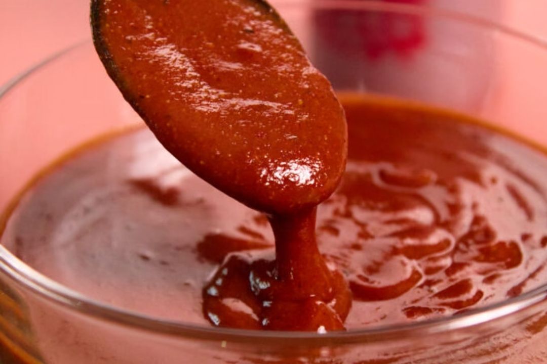 A spoon pours thick, red sauce into a glass bowl against a pink background.