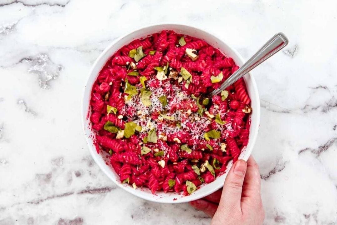 A hand holds a bowl of bright pink rotini pasta, topped with grated cheese, chopped pistachios, and herbs, with a fork placed inside the bowl on a marble surface.