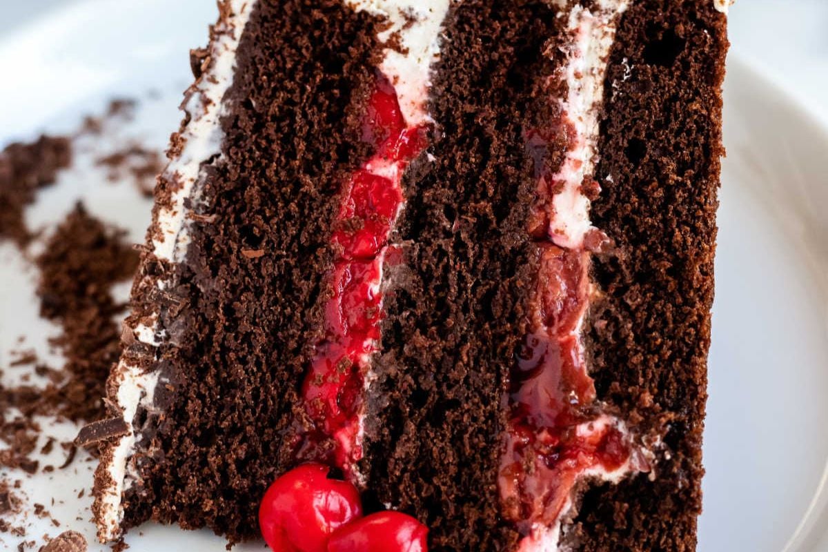 A close-up of a slice of chocolate cake with white frosting, cherry filling, chocolate shavings, and whole cherries on a white plate.