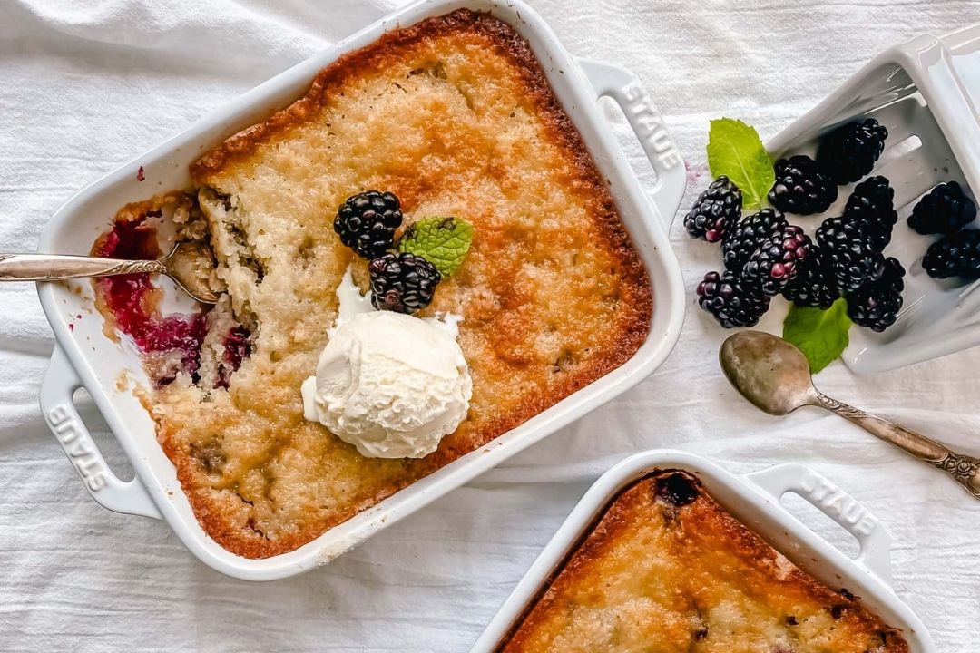 A baked cobbler in a white dish topped with a scoop of vanilla ice cream, blackberries, and a mint leaf. A spoon rests in the dish with a serving removed. Blackberries and a spoon are nearby.