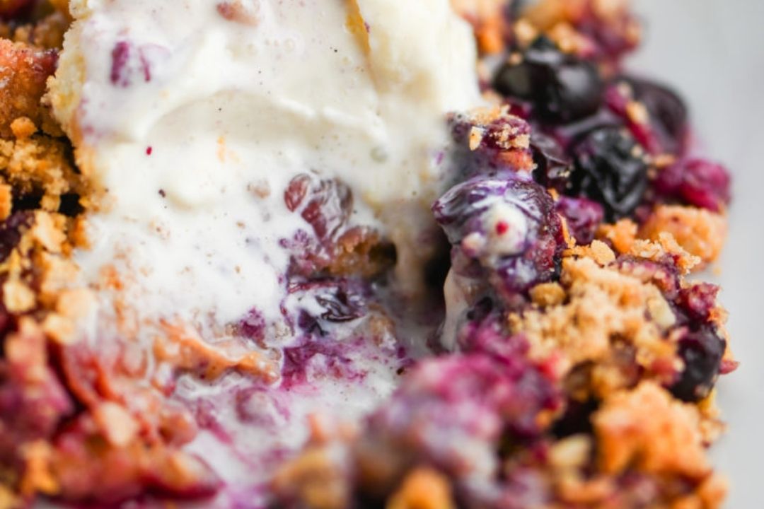 Close-up of a blueberry crumble dessert with a scoop of melting vanilla ice cream on top, showing crumbs, berries, and creamy texture.