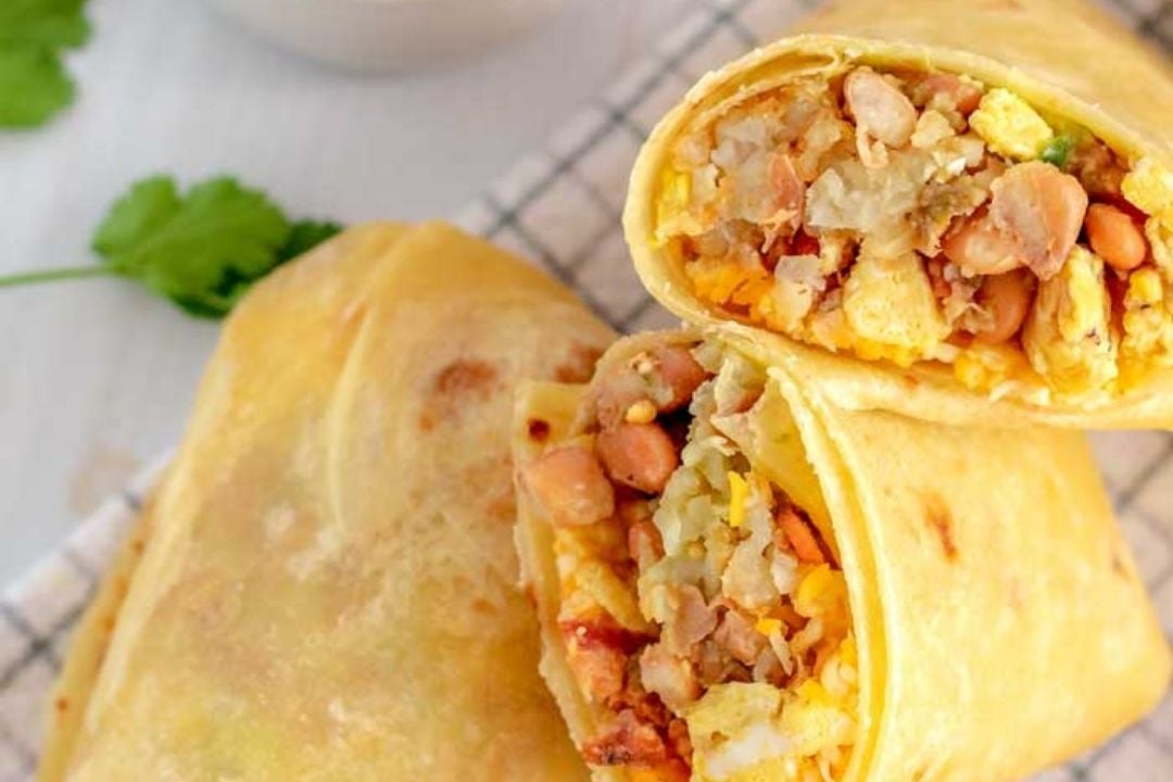 Close-up of a breakfast burrito cut in half, showing a filling of scrambled eggs, beans, potatoes, and cheese, placed on a checkered cloth with cilantro garnish nearby.