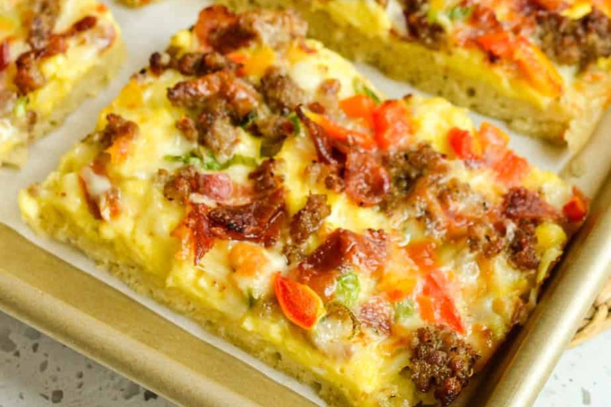 A rectangular breakfast pizza topped with sausage, bacon, melted cheese, and diced red and green peppers on a baking sheet.