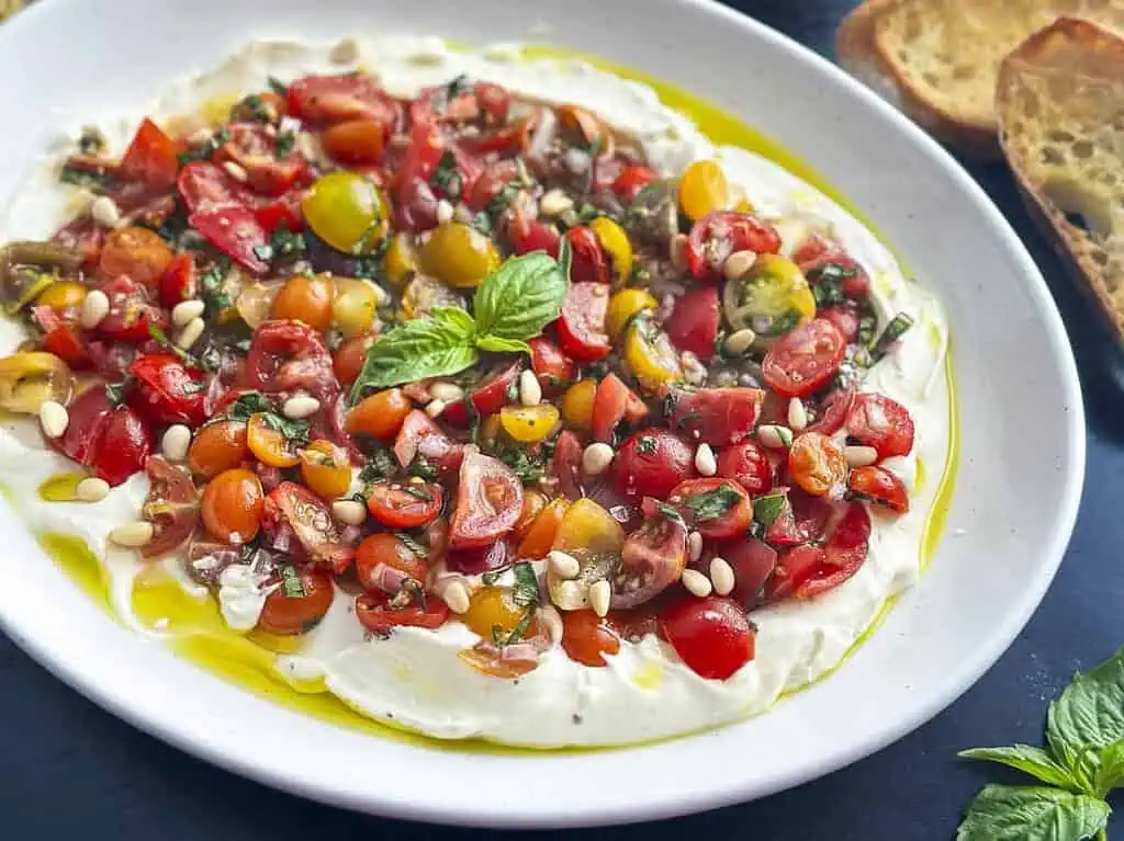 A plate of whipped ricotta topped with chopped cherry tomatoes, basil, pine nuts, and olive oil, served with toasted bread on the side.
