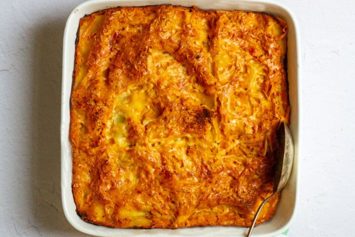 Baked cheesy casserole in a square white dish with a serving spoon on the side, showing a golden-brown crust and melted cheese.
