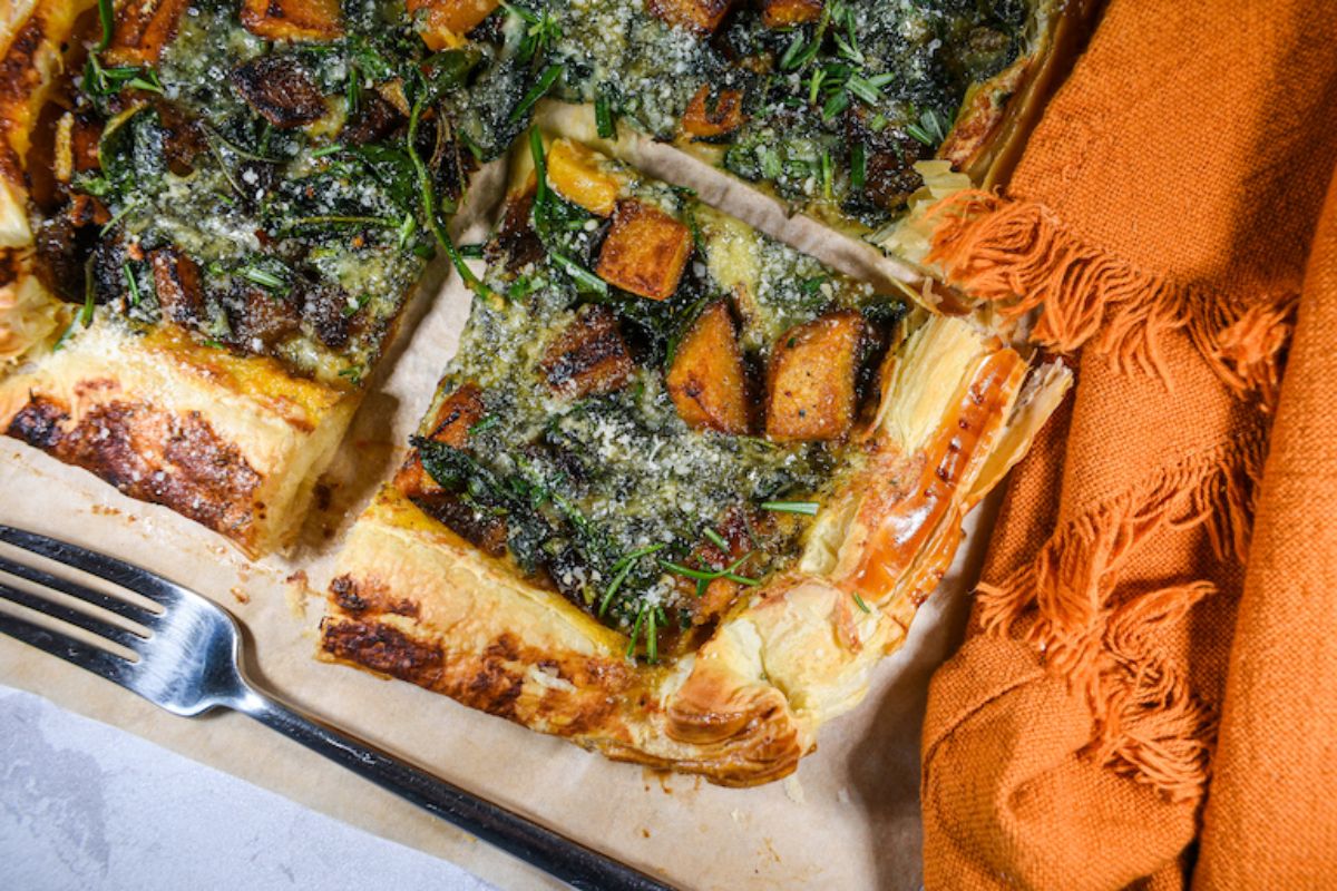 A baked puff pastry tart topped with roasted vegetables, herbs, and cheese, placed on parchment paper next to an orange cloth and a fork.