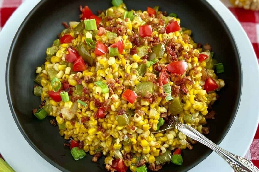 A black bowl filled with sautéed corn, diced red and green peppers, chopped green onions, tomatoes, and crumbled bacon, with a spoon resting inside.