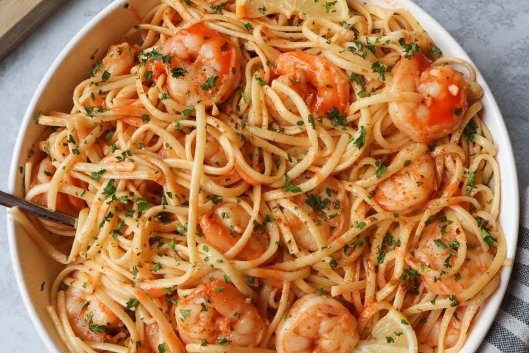 A bowl of linguine pasta mixed with shrimp, tomato-based sauce, lemon slices, and chopped parsley.