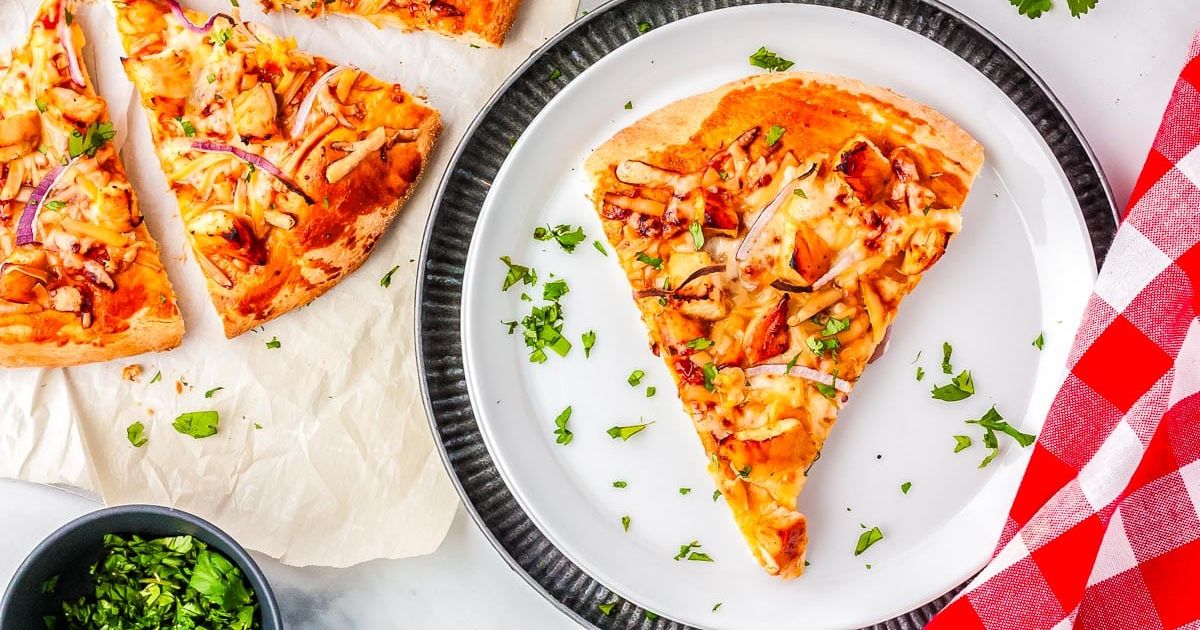 A slice of pizza with cheese, chicken, and red onion sits on a white plate, with more pizza and herbs nearby on a table with a red checkered napkin.