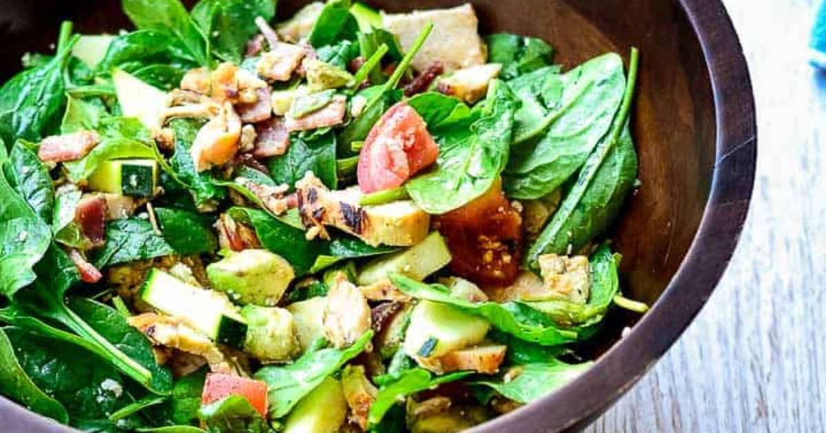 A wooden bowl filled with a spinach salad containing avocado, tomato, cucumber, zucchini, and chopped bacon pieces.