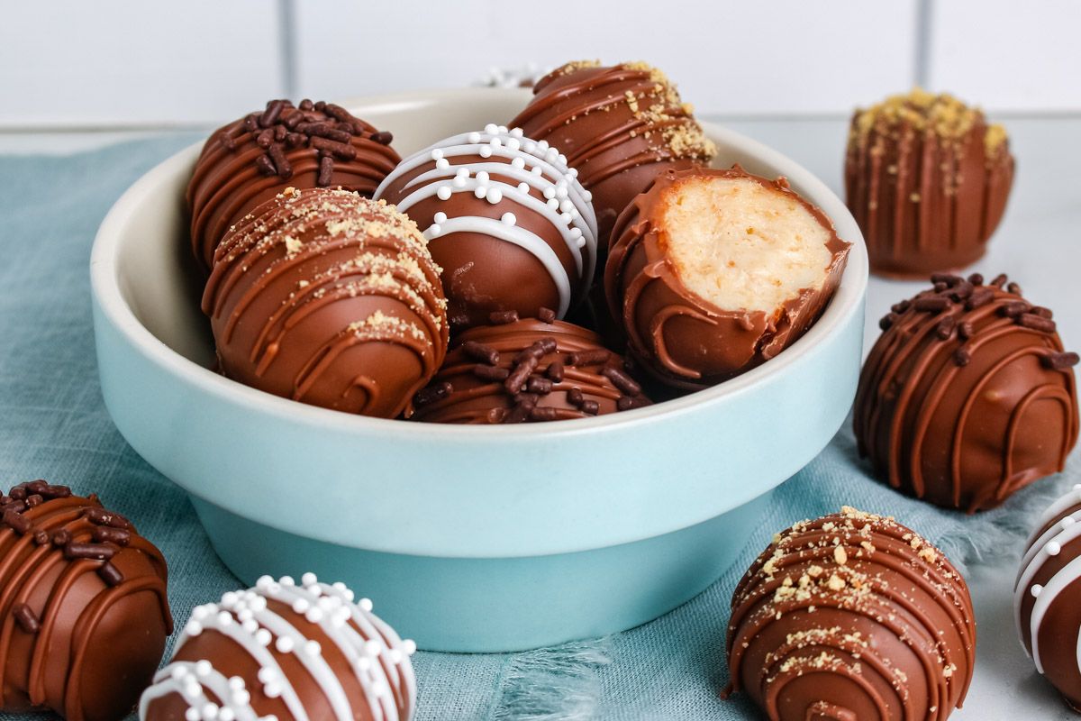 A bowl filled with chocolate-covered truffles, some decorated with sprinkles and white icing, with one truffle partially eaten to show the creamy filling inside.