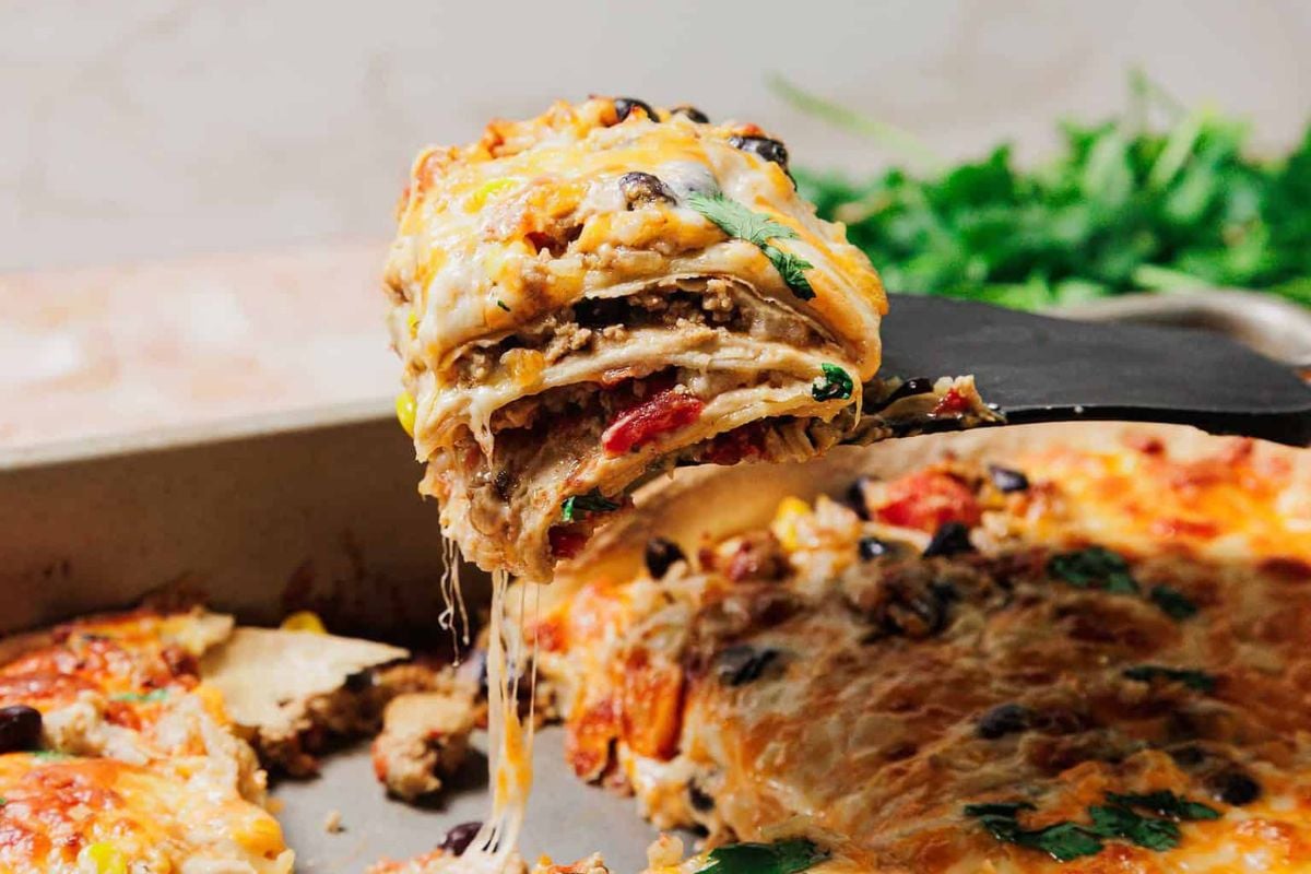 A spatula lifts a cheesy slice of layered casserole with visible vegetables, ground meat, and tortillas from a baking dish.