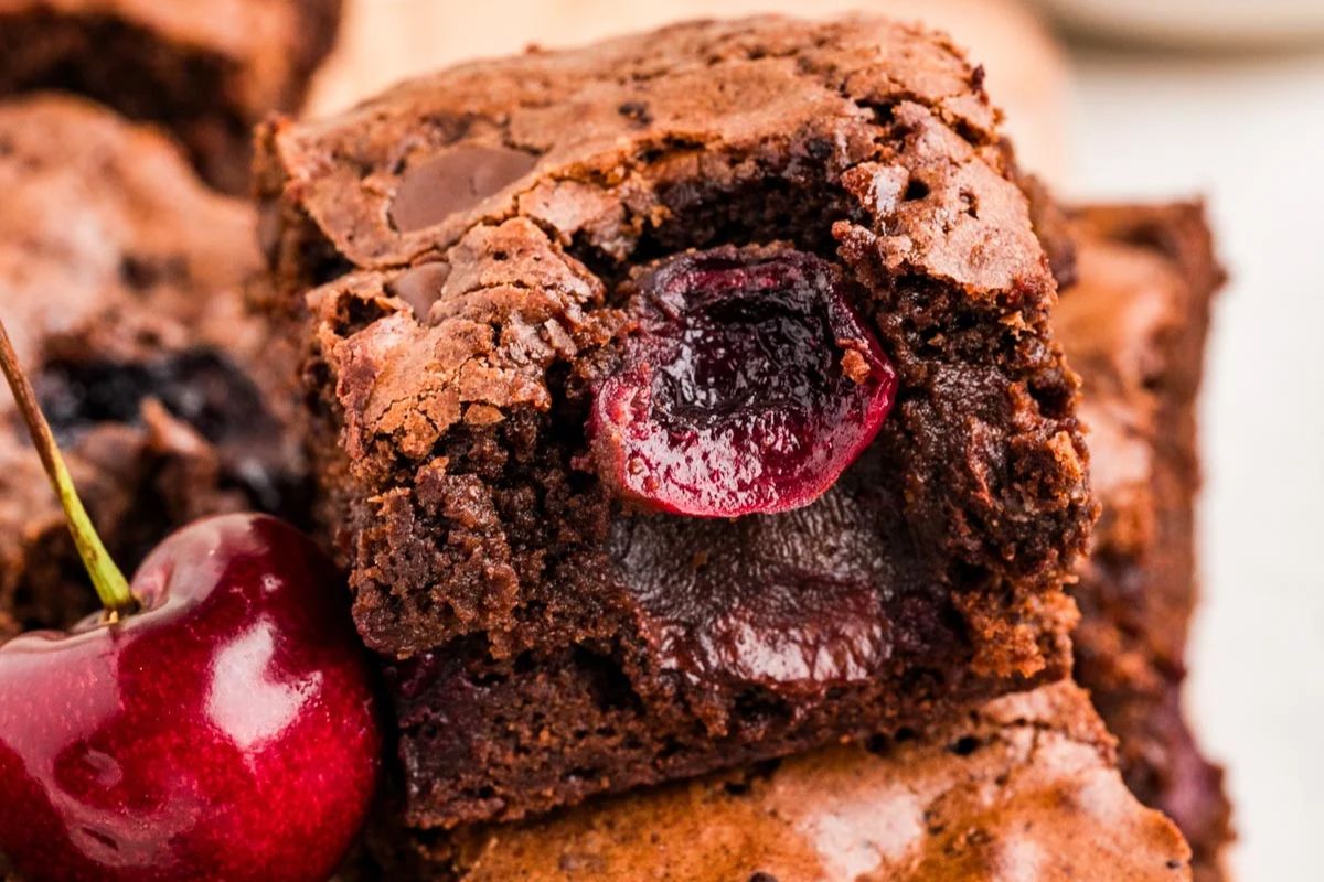Close-up of a chocolate brownie with a cherry inside, topped with a whole cherry beside it. The brownie has a crackled surface and moist interior.