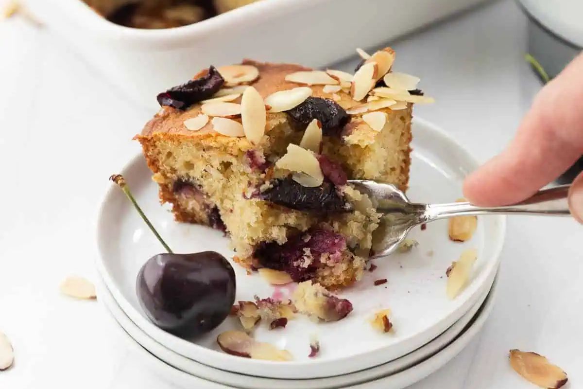 A hand holding a fork takes a bite of almond-topped cherry cake on a plate, with a fresh cherry and almond slices nearby.