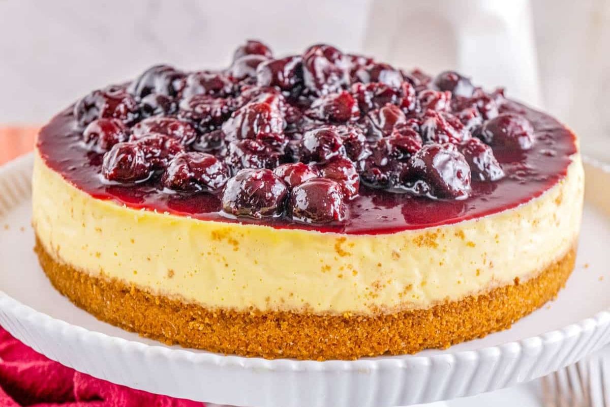 A cheesecake with a graham cracker crust, topped with a glossy layer of cherry compote, sits on a white cake stand.