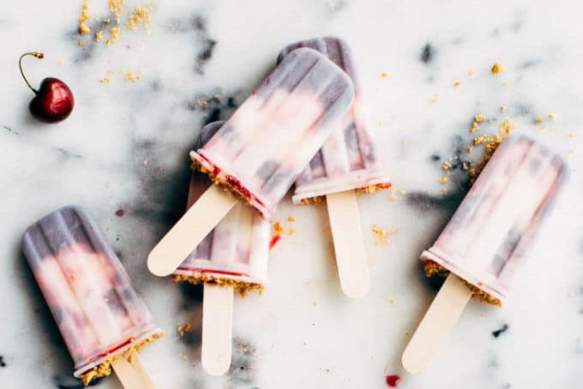 Five cherry swirl popsicles with wooden sticks are arranged on a marble surface; crumbs and a single cherry are scattered nearby.