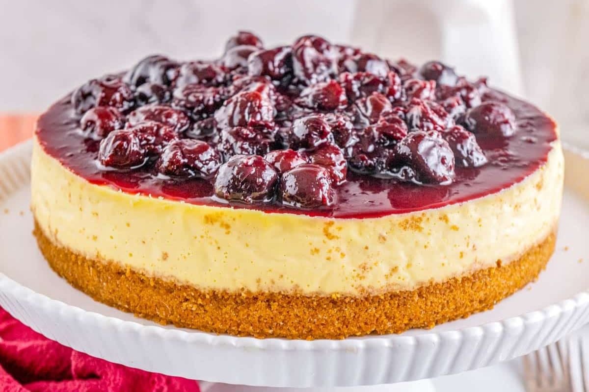 A cheesecake with a graham cracker crust and a cherry topping sits on a white cake stand.