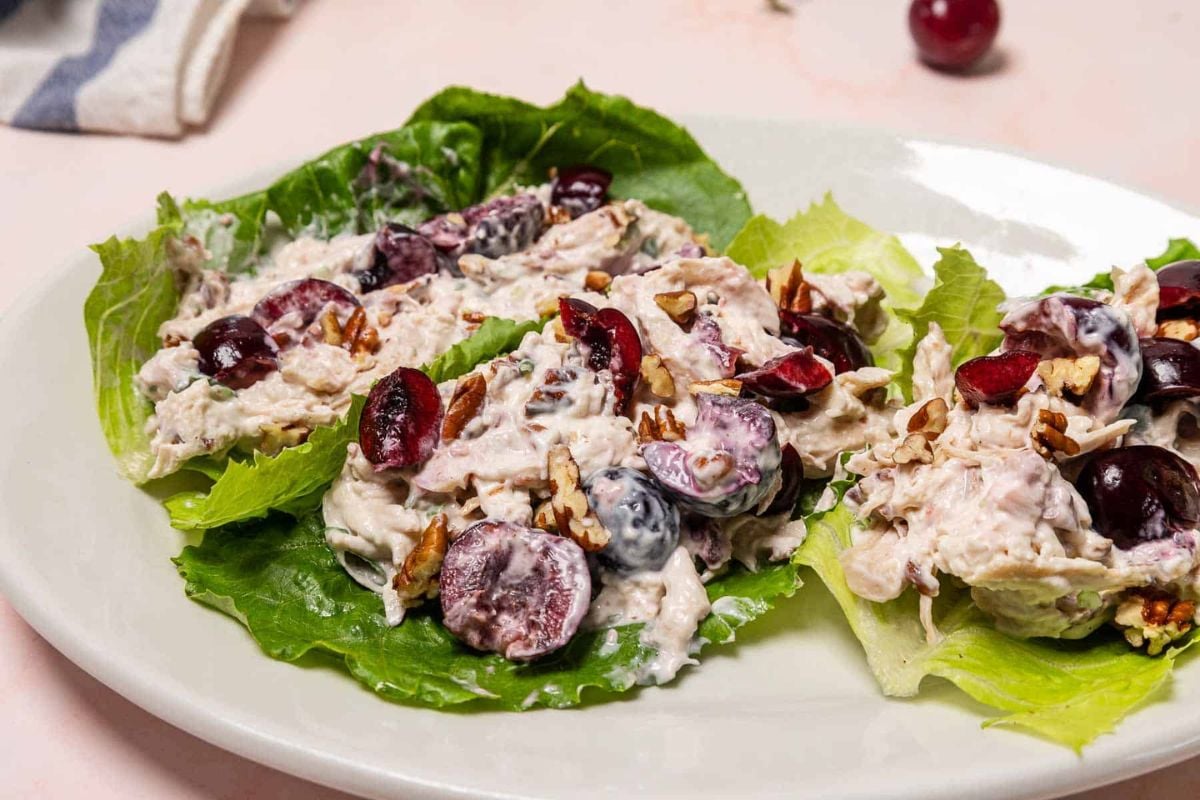 Three lettuce leaves topped with a creamy chicken salad containing halved grapes and chopped pecans, served on a white plate.