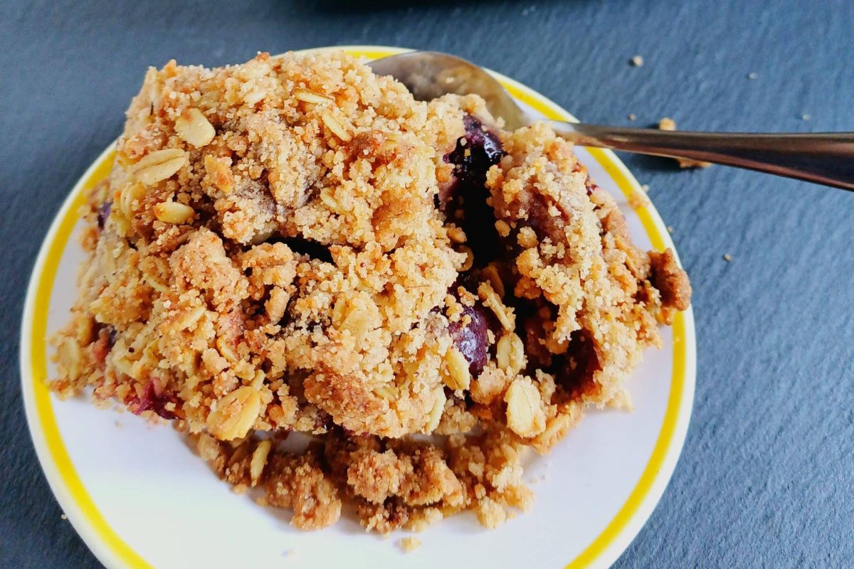 A serving of fruit crumble with oats on a white plate with a yellow rim, next to a metal spoon.