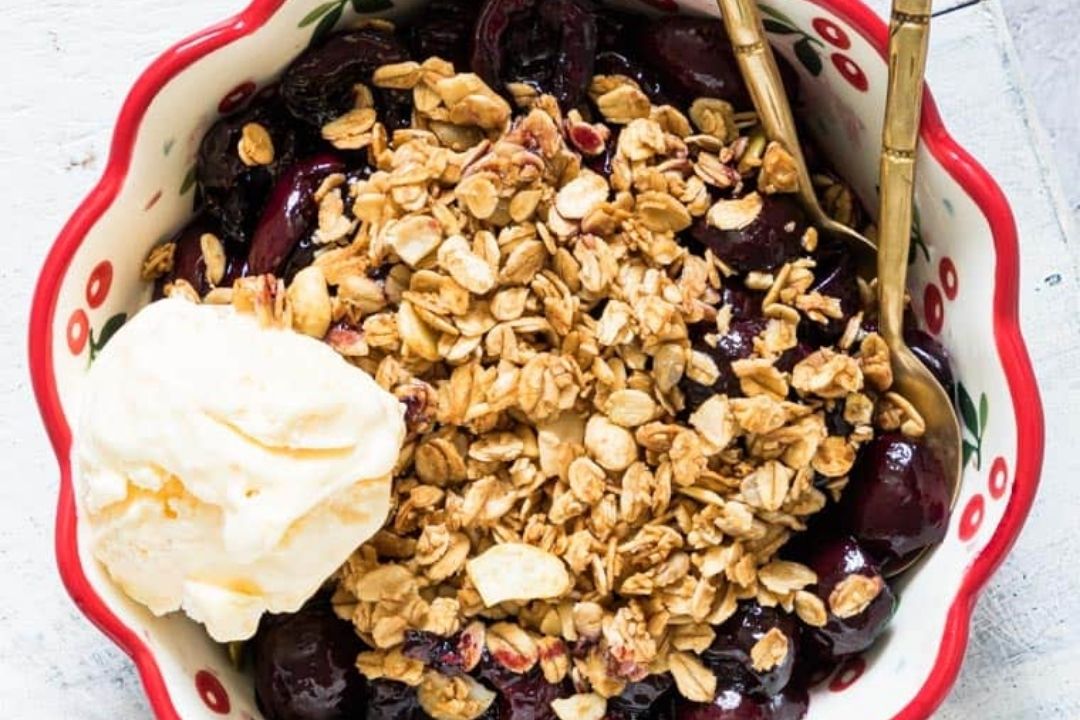 A bowl of cherry crisp topped with granola and a scoop of vanilla ice cream, served with two golden spoons.