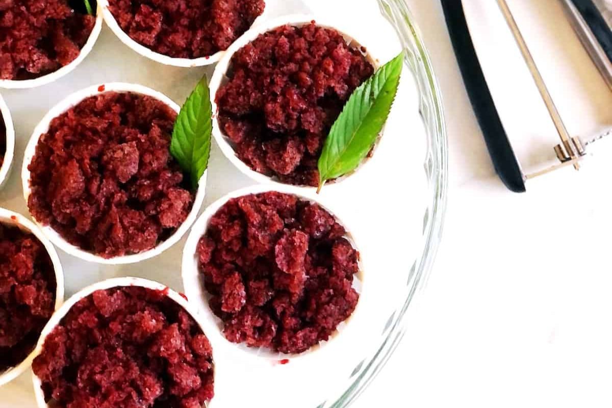 Cups of red granita topped with mint leaves arranged on a glass plate, with an ice cream scoop nearby.