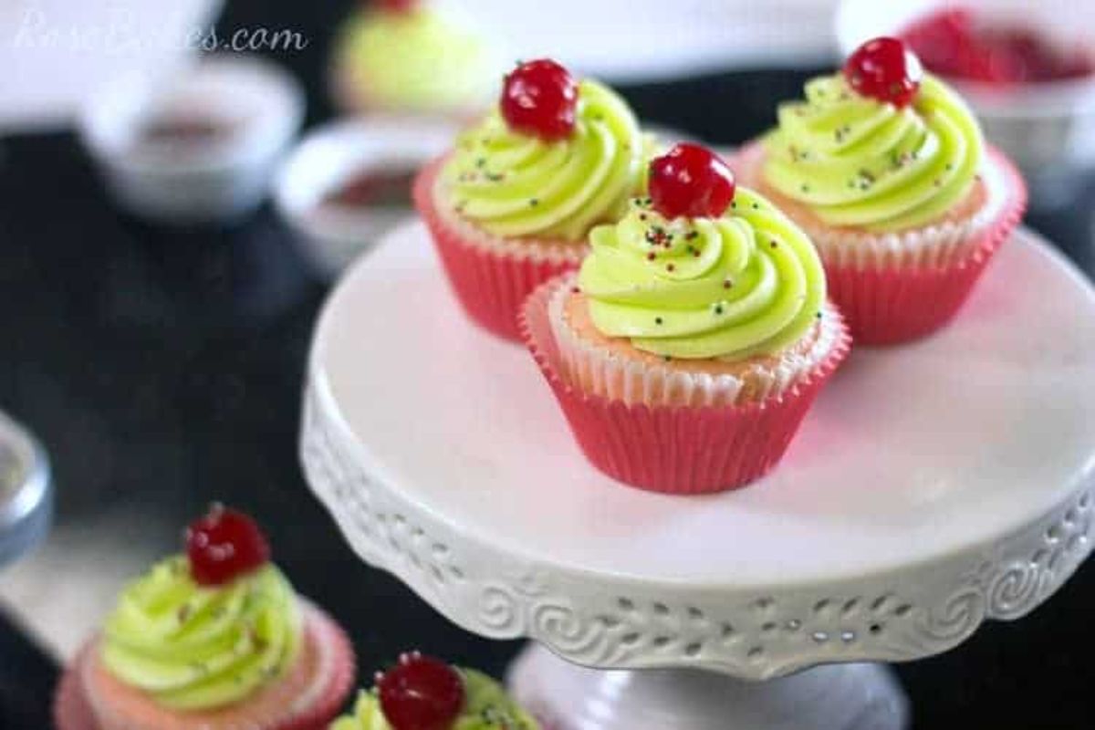 Three cupcakes with green frosting and cherries on top are displayed on a white cake stand, with more cupcakes and bowls in the background.