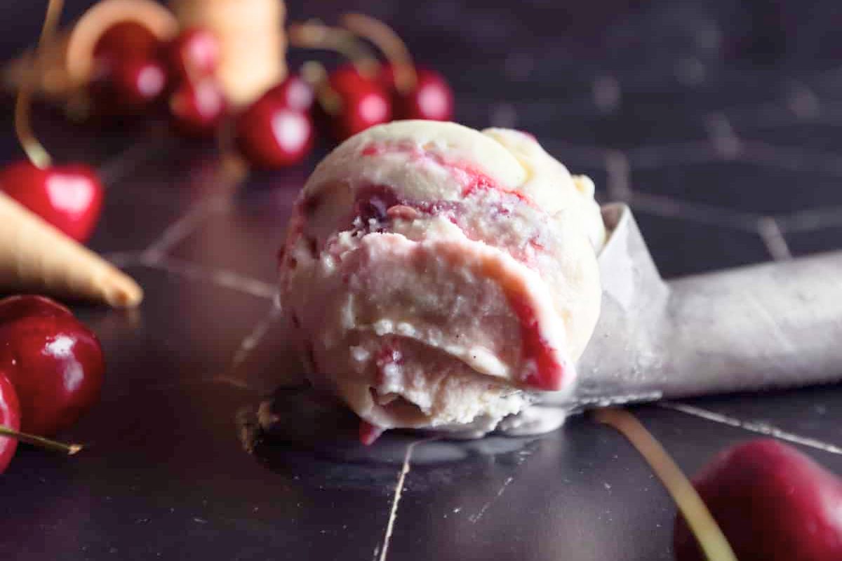 A scoop of cherry ice cream with visible cherry swirls sits on a dark surface, surrounded by fresh cherries and an ice cream cone in the background.