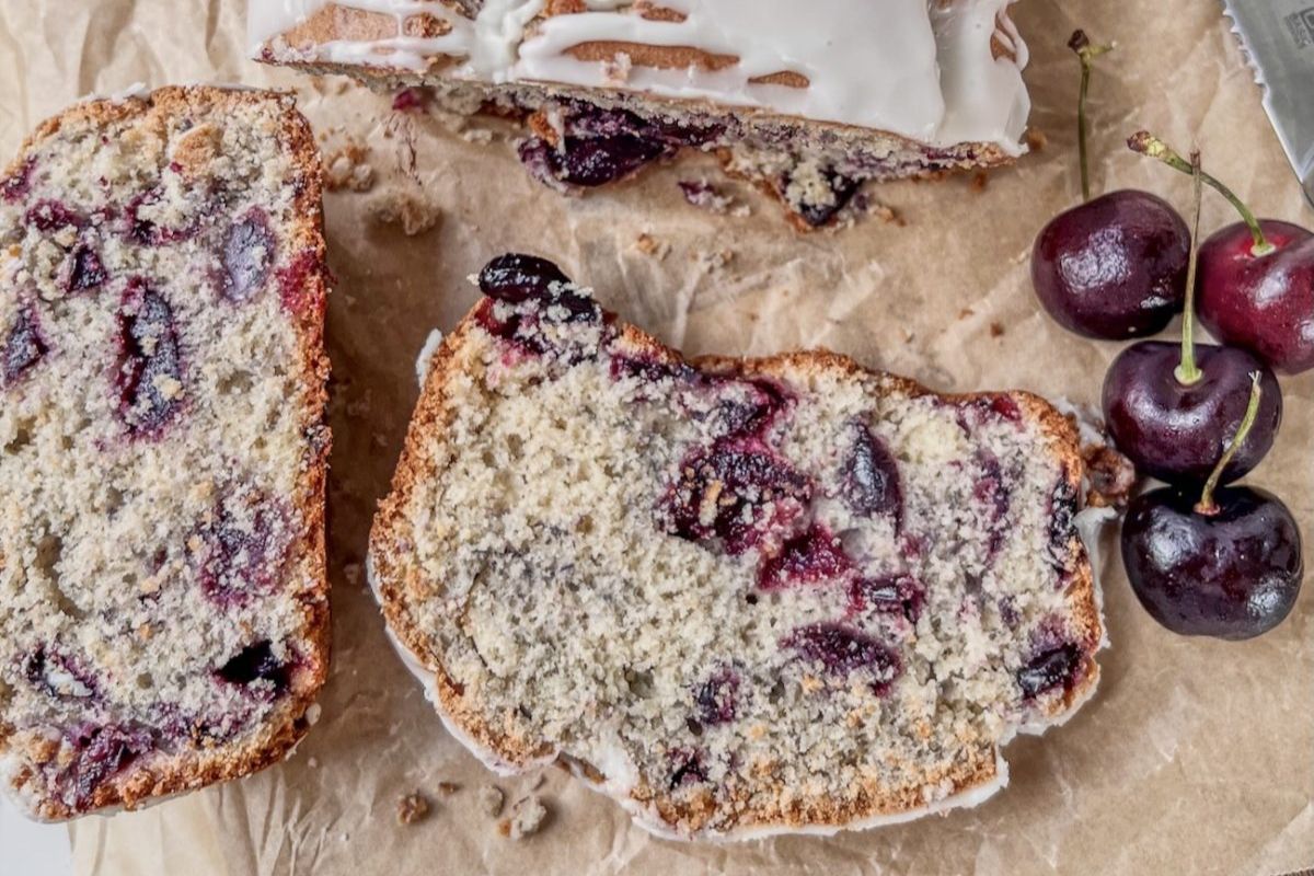 Sliced loaf of cherry bread with white icing on parchment paper, next to three fresh cherries.