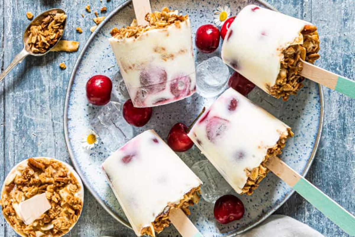 Four homemade yogurt and cherry popsicles with granola at the base are arranged on a plate with ice cubes and fresh cherries, with a spoonful of granola nearby.