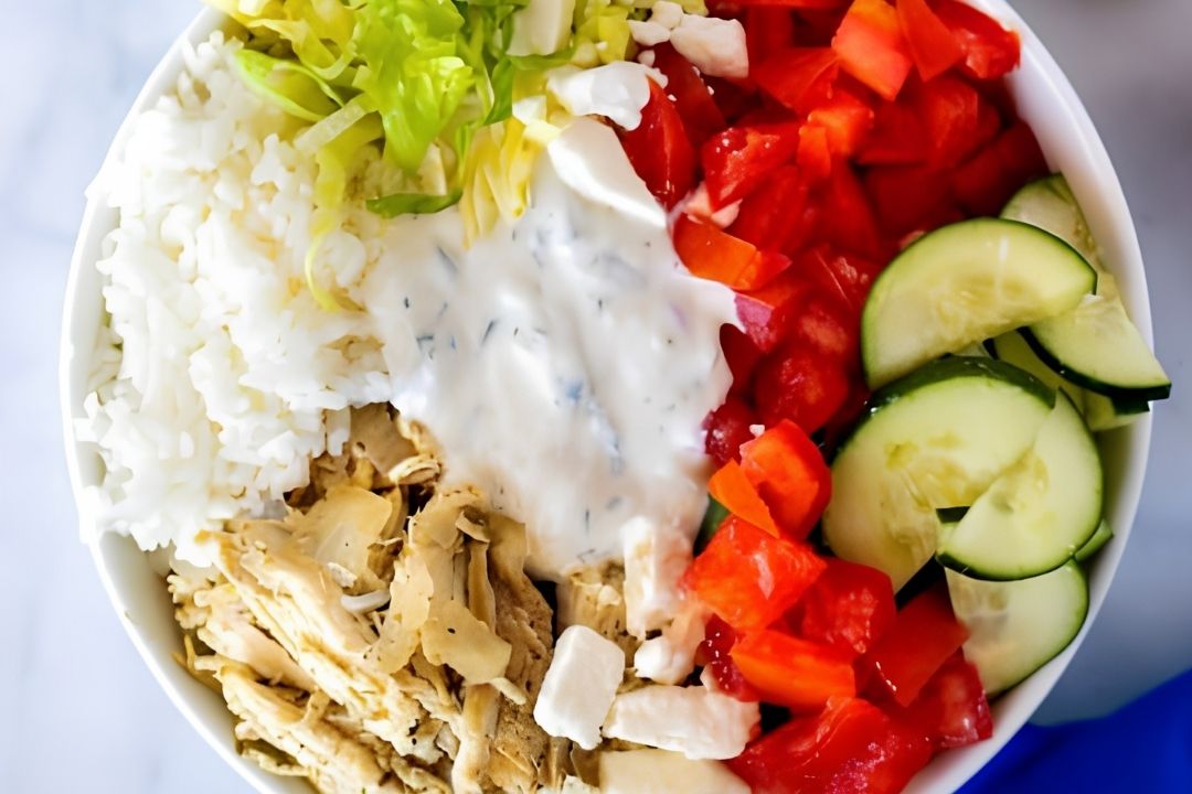 A bowl containing shredded chicken, white rice, chopped lettuce, diced tomatoes, cucumber slices, feta cheese, and a serving of creamy white dressing in the center.