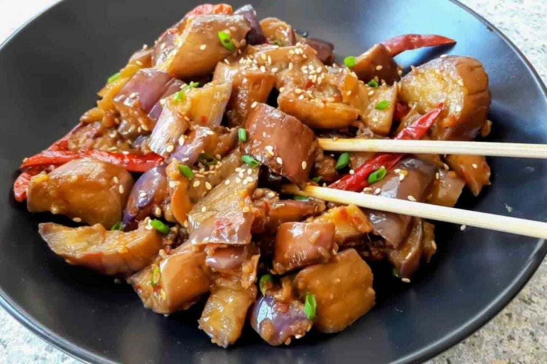 A black plate of stir-fried eggplant with red chilies, green onions, and sesame seeds, with chopsticks resting on the side.