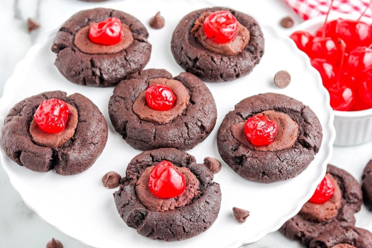 Six chocolate cookies topped with chocolate filling and a red cherry are arranged on a white plate, with extra cherries and chocolate chips nearby.