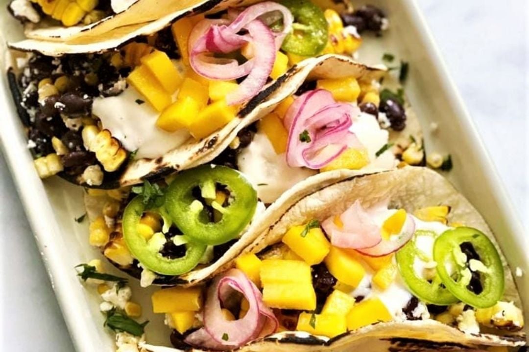 Three soft tacos filled with black beans, corn, mango, sour cream, sliced jalapeños, pickled onions, and cilantro on a rectangular white plate.