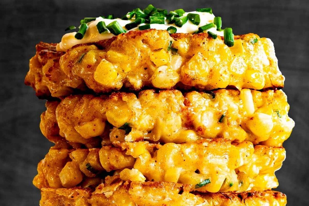 A stack of golden corn fritters topped with a dollop of sour cream and chopped chives against a dark background.