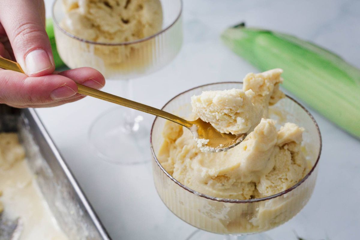 A hand holds a gold spoon scooping yellow ice cream from a glass dish, with another dish and an ear of corn in the background.