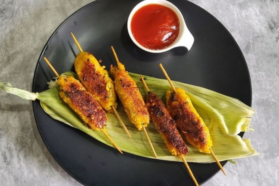 Five grilled skewers are arranged on a corn husk atop a black plate, with a small white bowl of red dipping sauce beside them.
