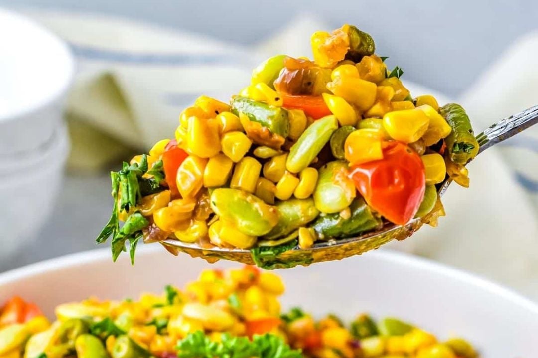 A close-up of a spoonful of succotash featuring corn, lima beans, tomatoes, and herbs, held above a bowl of the same dish.