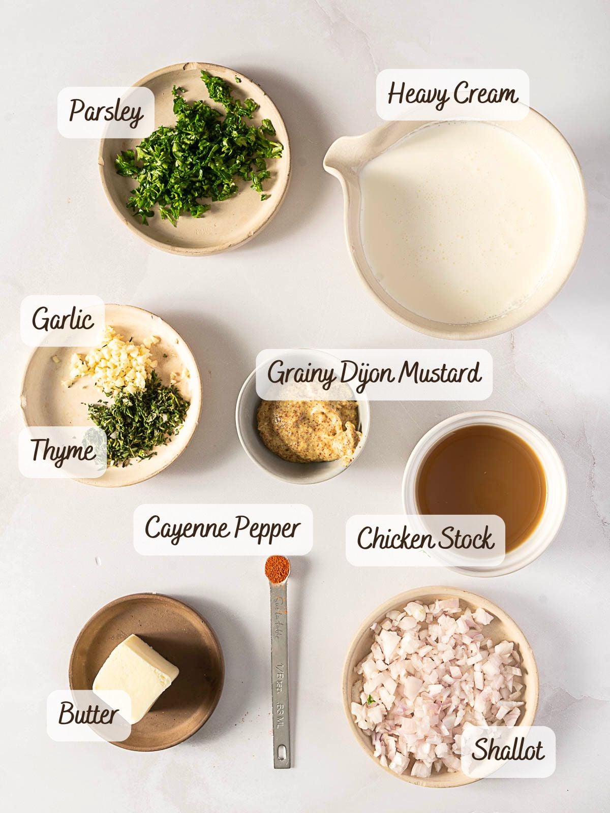 Nine small dishes with labeled ingredients—parsley, heavy cream, garlic, thyme, grainy Dijon mustard, chicken stock, cayenne pepper, butter, and shallot—are arranged on a white surface, showcasing everything you need to create a flavorful dijon cream sauce.