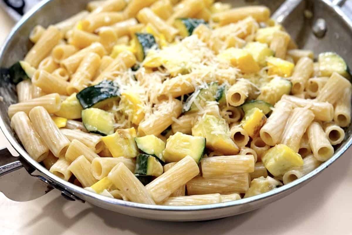 Cooked rigatoni pasta with diced zucchini and yellow squash, topped with shredded cheese, served in a metal skillet.